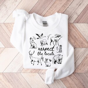 May include: White sweatshirt with a black graphic of various woodland animals, including a wolf, bear, and deer. The text "respect the locals" is printed in a handwritten style. The sweatshirt is folded on a wooden surface.
