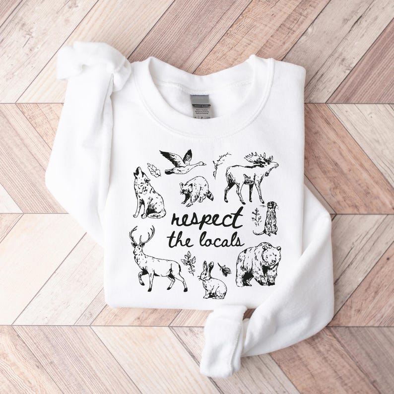 May include: White sweatshirt with a black graphic of various woodland animals, including a wolf, bear, and deer. The text "respect the locals" is printed in a handwritten style. The sweatshirt is folded on a wooden surface.