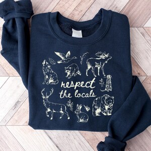 May include: Navy blue sweatshirt featuring a cream-colored graphic of various animals, including a wolf, moose, and bear. The text "respect the locals" is centered below the illustrations. The sweatshirt is folded on a wooden surface.