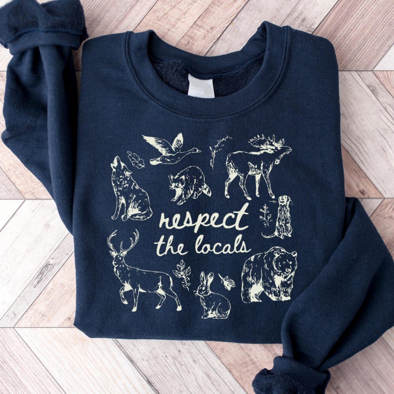 May include: Navy blue sweatshirt featuring a cream-colored graphic of various animals, including a wolf, moose, and bear. The text "respect the locals" is centered below the illustrations. The sweatshirt is folded on a wooden surface.