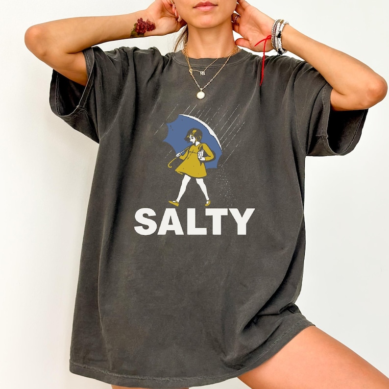 May include: Dark grey t-shirt featuring a cartoon illustration of a person holding a blue umbrella in the rain. The person is wearing a yellow dress. The word "SALTY" is printed in white block letters below the illustration.
