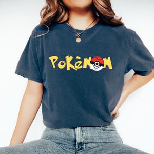 Puede incluir: Una camiseta azul marino con la palabra "Pok&eacute;mon" en amarillo y un gr&aacute;fico de Pok&eacute; Ball rojo y blanco. La camiseta tiene mangas cortas y cuello redondo.