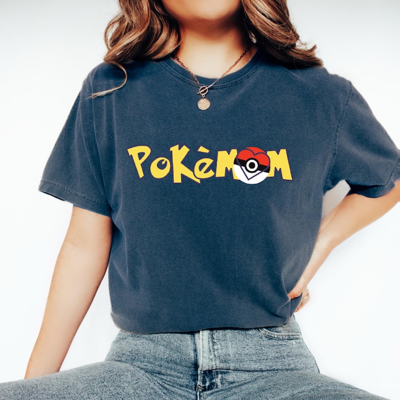 Puede incluir: Una camiseta azul marino con la palabra "Pok&eacute;mon" en amarillo y un gr&aacute;fico de Pok&eacute; Ball rojo y blanco. La camiseta tiene mangas cortas y cuello redondo.