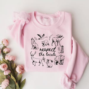 May include: A light pink sweatshirt with a crew neck and long sleeves. The front features a black graphic of various animals, including a wolf, bear, and deer, with the text "respect the locals."