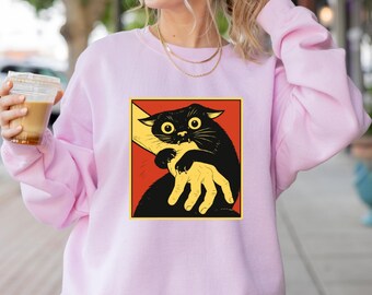 May include: A light pink sweatshirt featuring a graphic of a black cat biting a yellow hand, set against a red background. The cat has wide eyes and is in a distressed pose. The sweatshirt is a solid color.
