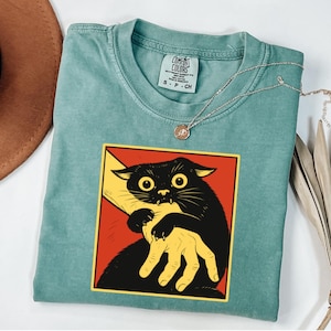 May include: A teal Comfort Colors t-shirt featuring a graphic of a black cat biting a yellow hand, set against a red background. The shirt has a crew neck and short sleeves. A silver necklace with a small pendant is draped across the shirt.