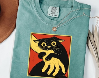 May include: A teal Comfort Colors t-shirt featuring a graphic of a black cat biting a yellow hand, set against a red background. The shirt has a crew neck and short sleeves. A silver necklace with a small pendant is draped across the shirt.