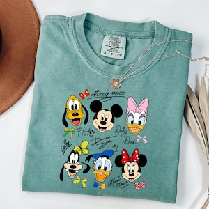 Peut inclure: Un t-shirt Comfort Colors vert d'eau avec un graphisme de Mickey Mouse et ses amis. Le motif comprend Pluto, Mickey, Daisy, Donald, Dingo et Minnie Mouse. Le t-shirt a un col rond et des manches courtes.