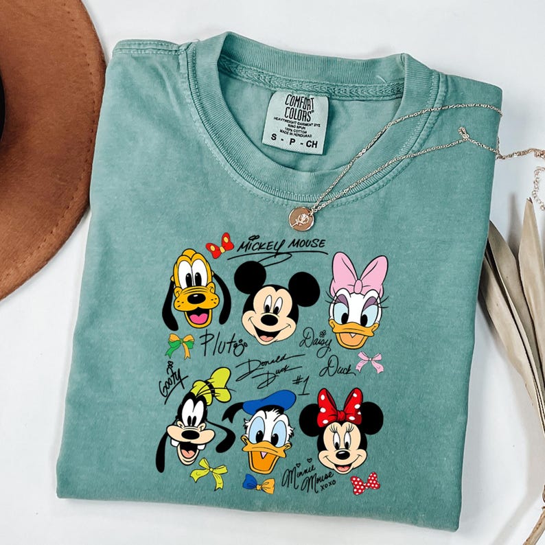 Peut inclure: Un t-shirt Comfort Colors vert d'eau avec un graphisme de Mickey Mouse et ses amis. Le motif comprend Pluto, Mickey, Daisy, Donald, Dingo et Minnie Mouse. Le t-shirt a un col rond et des manches courtes.
