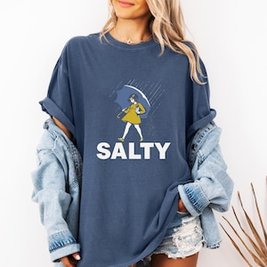 May include: Navy blue t-shirt with a graphic of a person holding an umbrella in the rain. The person is wearing a yellow dress. The word "SALTY" is printed in white block letters below the graphic.
