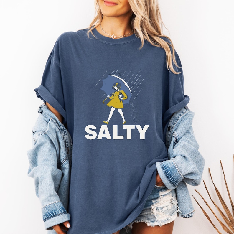 May include: Navy blue t-shirt with a graphic of a person holding an umbrella in the rain. The person is wearing a yellow dress. The word "SALTY" is printed in white block letters below the graphic.