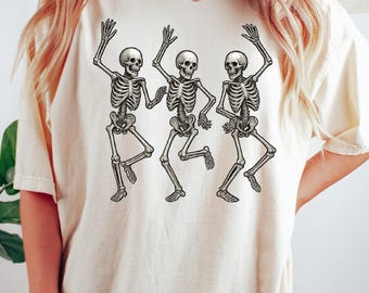 May include: Cream-colored t-shirt featuring a graphic of three dancing skeletons. The skeletons are depicted in black and white, with their arms raised in a celebratory pose. The design is centered on the front of the shirt.