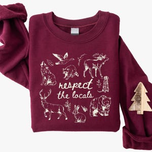 May include: Burgundy sweatshirt with a white graphic of various animals and the text "respect the locals." The design includes a wolf, raccoon, moose, bear, deer, rabbit, and other wildlife. A wooden tree-shaped pin is attached.