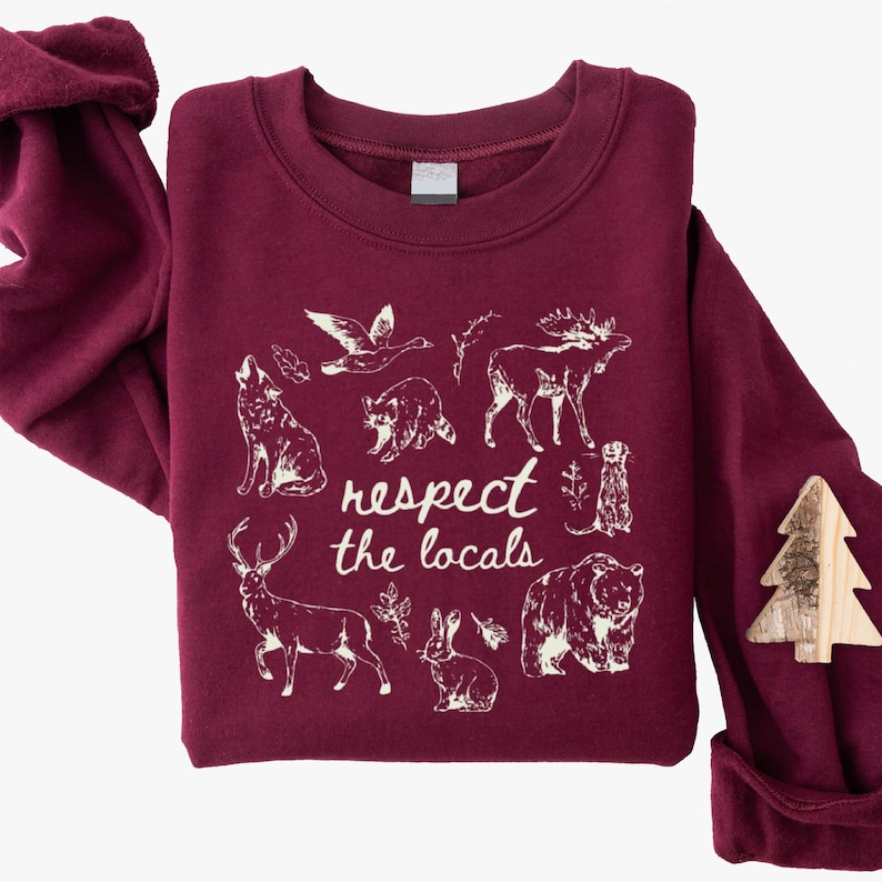 May include: Burgundy sweatshirt with a white graphic of various animals and the text "respect the locals." The design includes a wolf, raccoon, moose, bear, deer, rabbit, and other wildlife. A wooden tree-shaped pin is attached.