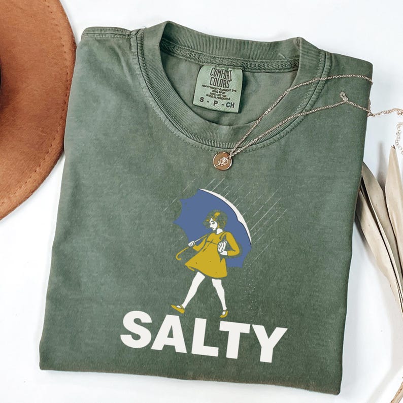 May include: Olive green t-shirt with the word "SALTY" in white block letters. The shirt features a graphic of a person in a yellow dress holding a blue umbrella in the rain. A gold necklace is draped across the shirt.