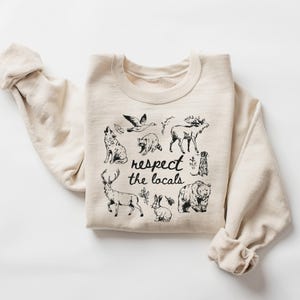 May include: A cream-colored sweatshirt featuring a black graphic of various woodland animals, including a wolf, bear, and deer. The phrase "respect the locals" is written in a handwritten style below the animal illustrations. The sleeves are rolled up.
