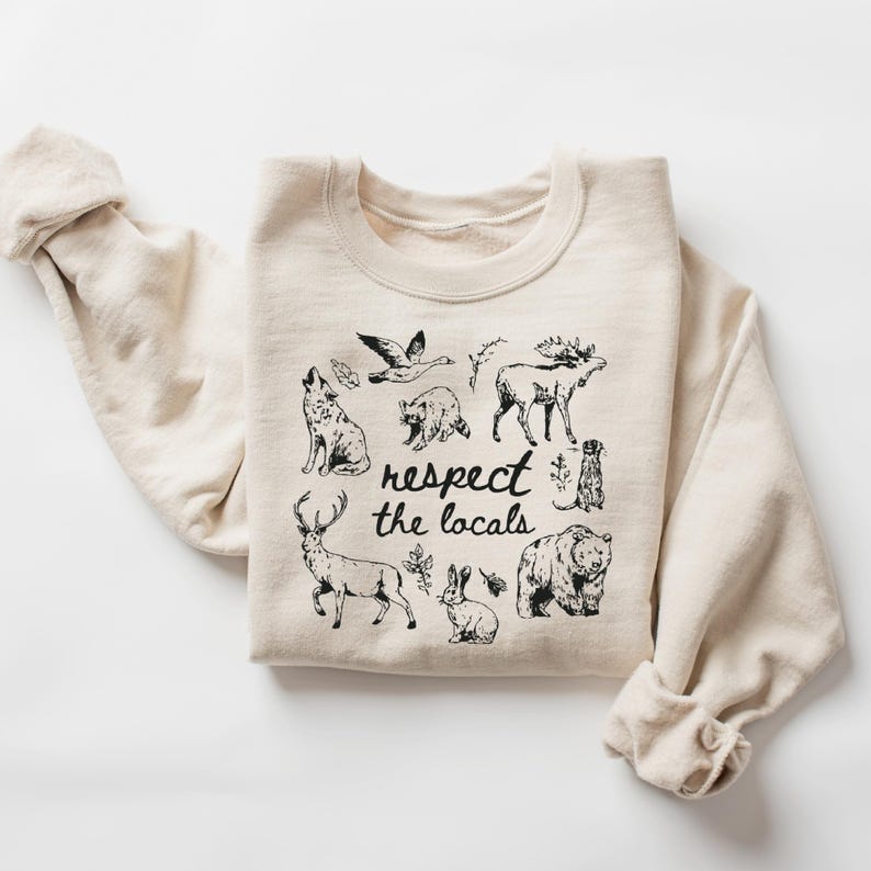 May include: A cream-colored sweatshirt featuring a black graphic of various woodland animals, including a wolf, bear, and deer. The phrase "respect the locals" is written in a handwritten style below the animal illustrations. The sleeves are rolled up.
