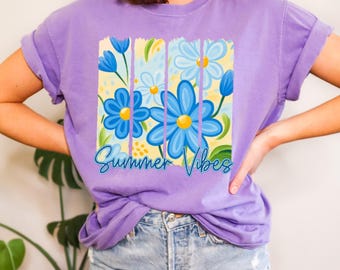 May include: A lilac-colored t-shirt featuring a floral design with blue flowers and yellow centers. The graphic includes the text "Summer Vibes" in a script font. The shirt has rolled-up sleeves and is paired with denim shorts.