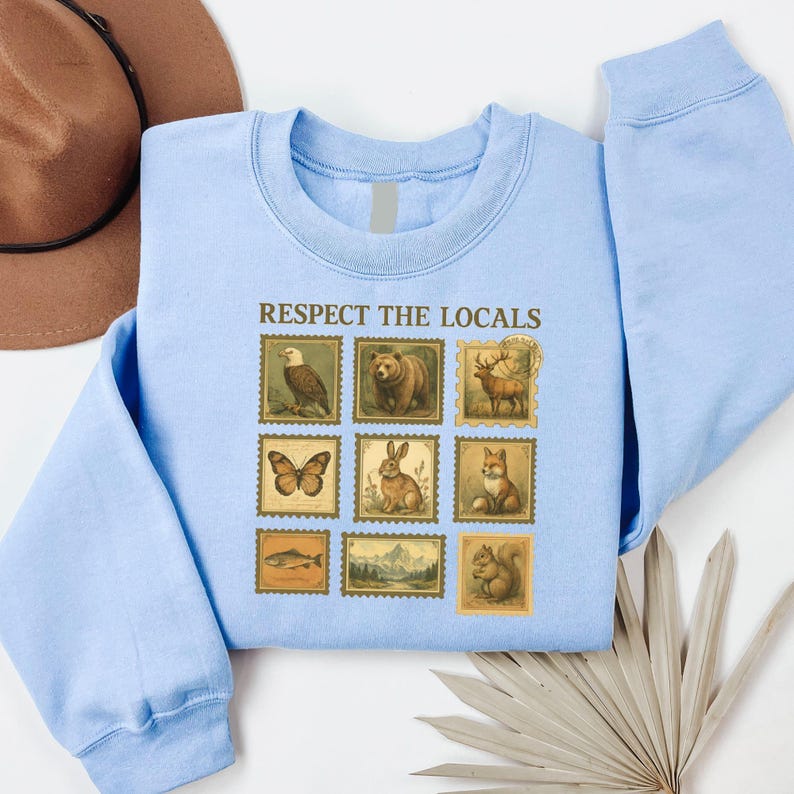 May include: Light blue sweatshirt with the phrase "RESPECT THE LOCALS" above nine postage stamp-style illustrations of wildlife. The stamps feature an eagle, bear, deer, butterfly, rabbit, fox, fish, mountain scene, and squirrel.