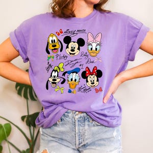 Peut inclure: Un t-shirt lilas avec un motif de Mickey Mouse et ses amis. Le motif comprend Pluto, Mickey, Daisy Duck, Dingo, Donald Duck et Minnie Mouse, avec leurs noms et signatures. Le t-shirt a des manches retrouss&eacute;es.