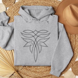 Puede incluir: Una sudadera con capucha gris claro con un dise&ntilde;o cosido en negro. El dise&ntilde;o es un patr&oacute;n abstracto y sim&eacute;trico. La sudadera con capucha tiene una capucha con cord&oacute;n y mangas largas. El fondo incluye una estera tejida y un sombrero marr&oacute;n.