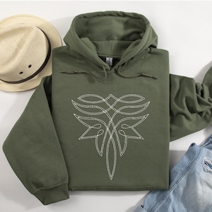 Puede incluir: Una sudadera con capucha verde oliva con un dise&ntilde;o floral blanco estilizado en la parte delantera. La sudadera tiene una capucha con cord&oacute;n y mangas largas. Un sombrero de paja y unos vaqueros tambi&eacute;n est&aacute;n en la imagen.
