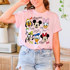 Peut inclure: T-shirt rose clair avec des visages de personnages de dessins anim&eacute;s, dont Mickey Mouse, Minnie Mouse, Donald Duck, Daisy Duck, Dingo et Pluto. Les mots "Mickey Mouse" sont &eacute;crits au-dessus des personnages.