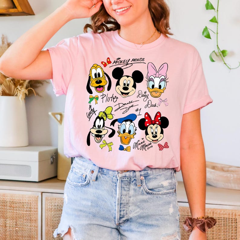Peut inclure: T-shirt rose clair avec des visages de personnages de dessins anim&eacute;s, dont Mickey Mouse, Minnie Mouse, Donald Duck, Daisy Duck, Dingo et Pluto. Les mots "Mickey Mouse" sont &eacute;crits au-dessus des personnages.