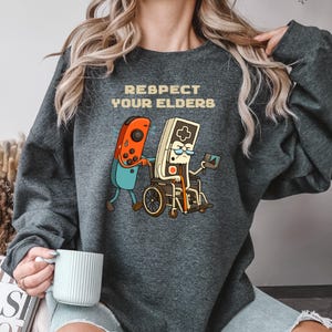 May include: Dark gray sweatshirt with the text "RESPECT YOUR ELDERS" above a cartoon graphic. The graphic depicts a red and blue video game controller pushing a wheelchair with a retro game controller inside. The retro controller is holding a small screen.