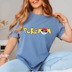 Puede incluir: Una camiseta azul con la palabra "Pok&eacute;mon" en letras amarillas y un dise&ntilde;o de Pok&eacute; Ball rojo y blanco. La camiseta de manga corta est&aacute; hecha de un material suave.