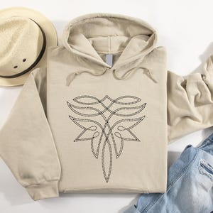 Puede incluir: Sudadera con capucha beige con un dise&ntilde;o floral abstracto negro cosido en la parte delantera. La sudadera tiene una capucha con cord&oacute;n y un bolsillo de canguro. Se ven un sombrero beige y una chaqueta vaquera.