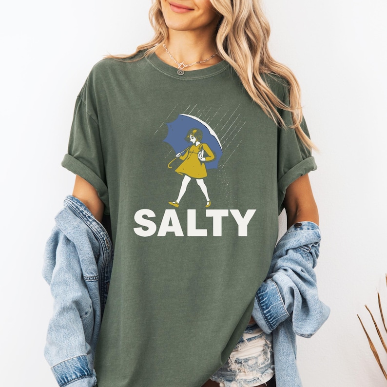 May include: Olive green t-shirt with the word "SALTY" in white block letters. The shirt features a graphic of a person in a yellow dress and blue umbrella walking in the rain. The shirt has rolled-up sleeves.