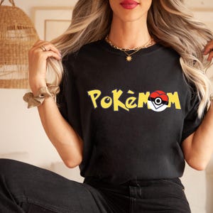 Puede incluir: Camiseta negra con la palabra "Pok&eacute;mon" en amarillo, con un dise&ntilde;o de Pok&eacute; Ball rojo, blanco y negro. Un estilo casual, adecuado para el uso diario.