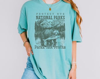 May include: A turquoise t-shirt with a graphic that reads "Protect Our National Parks" above an illustration of a bear holding a fish in a lake. The text "Parks, Not Profits" is below the image.