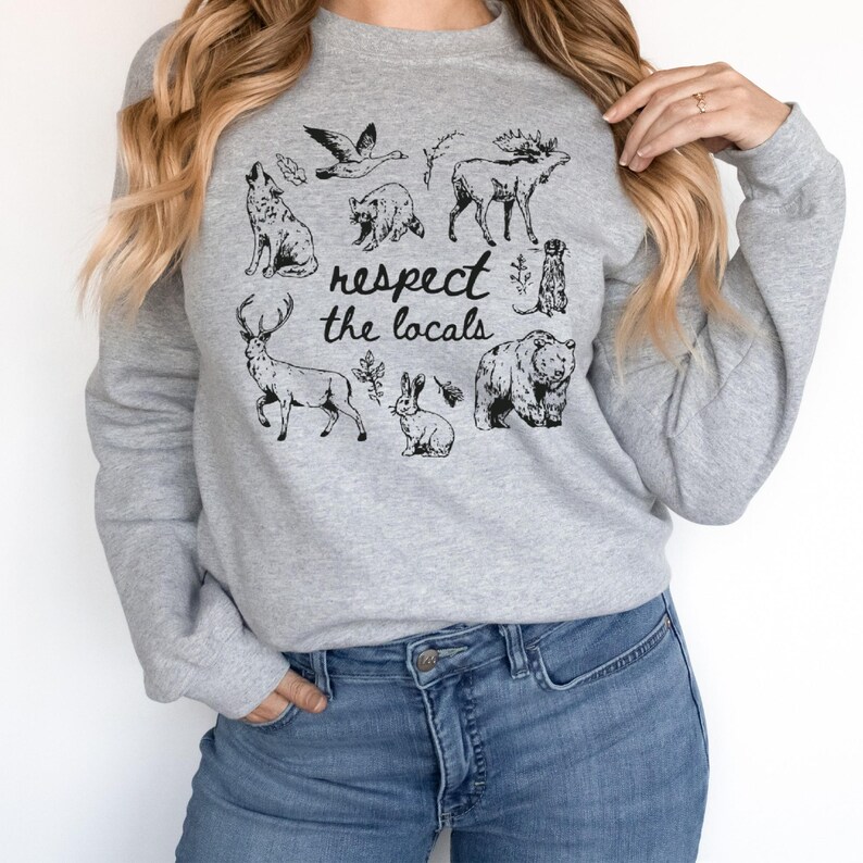 May include: A light gray sweatshirt featuring a collection of black and white illustrations of various animals, including a wolf, bear, and deer. The phrase "respect the locals" is printed in a handwritten style font.