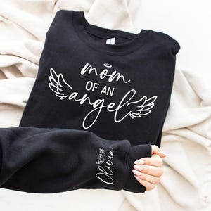 May include: Black sweatshirt with white text that reads "mom of an angel" with angel wings on either side. The left sleeve has a small white angel wing with the name "Olivia" underneath.