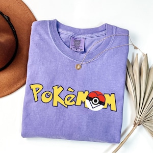 Puede incluir: Una camiseta de color lila con la palabra "Pok&eacute;mon" en letras amarillas y un dise&ntilde;o de coraz&oacute;n Pok&eacute; Ball rojo, blanco y negro. La camiseta tiene cuello redondo y mangas cortas. Tambi&eacute;n se ven un collar dorado y un sombrero marr&oacute;n.