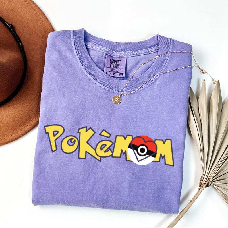 Puede incluir: Una camiseta de color lila con la palabra "Pok&eacute;mon" en letras amarillas y un dise&ntilde;o de coraz&oacute;n Pok&eacute; Ball rojo, blanco y negro. La camiseta tiene cuello redondo y mangas cortas. Tambi&eacute;n se ven un collar dorado y un sombrero marr&oacute;n.