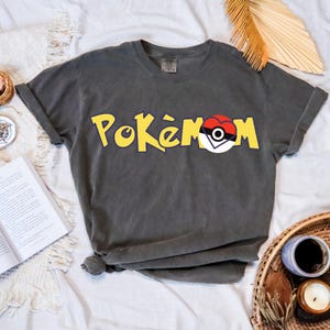 Puede incluir: Camiseta gris oscuro con la palabra "Pok&eacute;mon" en amarillo y un dise&ntilde;o de Pok&eacute; Ball rojo, blanco y negro. La camiseta es de material suave y tiene las mangas enrolladas.
