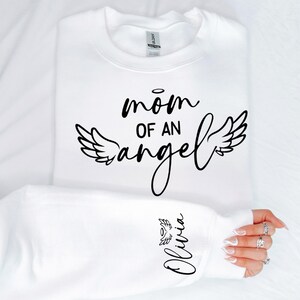 May include: White sweatshirt with black text that reads "mom of an angel" with angel wings on either side. The left sleeve has the name "Olivia" written in black with angel wings.