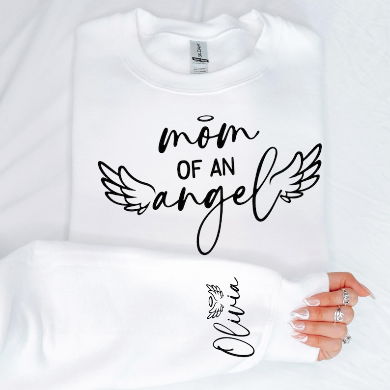 May include: White sweatshirt with black text that reads "mom of an angel" with angel wings on either side. The left sleeve has the name "Olivia" written in black with angel wings.