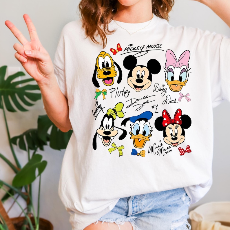 Peut inclure: T-shirt blanc avec un motif color&eacute; de Mickey Mouse, Minnie Mouse, Donald Duck, Daisy Duck, Dingo et Pluto. Le motif comprend les visages et les signatures des personnages. Les mots "Mickey Mouse" sont &eacute;crits en haut.