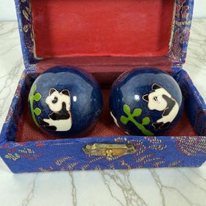 May include: A pair of blue Chinese health balls with panda designs, nestled in a blue and red patterned box. The balls feature black and white panda illustrations with green accents. The box has a gold clasp.