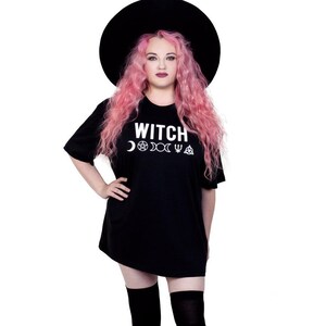 May include: Black t-shirt with the word "WITCH" in white, along with crescent moon, pentagram, and other symbols. The person in the image is wearing a black hat and has pink hair.
