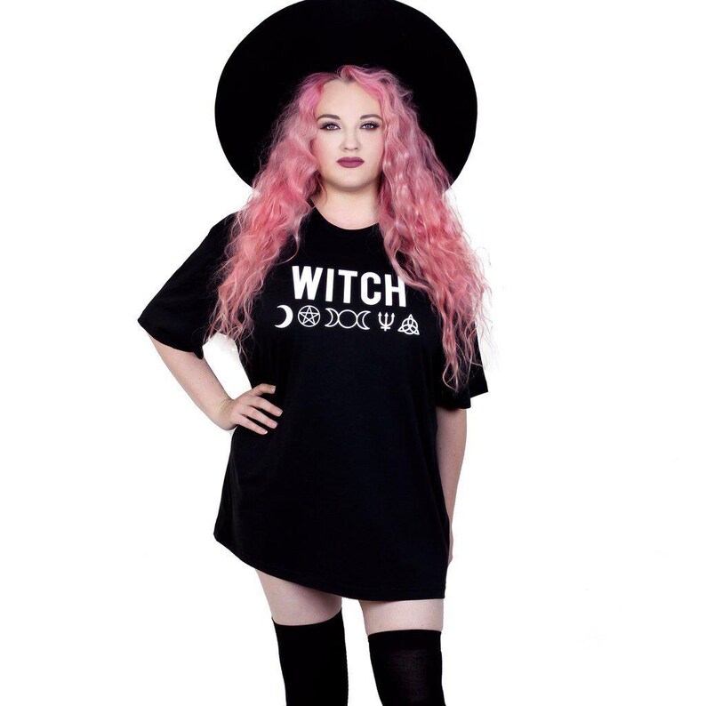 May include: Black t-shirt with the word "WITCH" in white, along with crescent moon, pentagram, and other symbols. The person in the image is wearing a black hat and has pink hair.