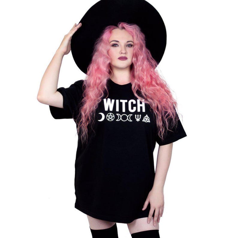 May include: Black t-shirt with the word "WITCH" in white, along with crescent moon, pentagram, and other symbols. The person in the image is wearing a large black hat and has pink hair.