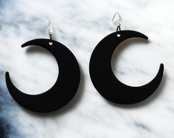 May include: A pair of black crescent moon earrings. The earrings are made of a solid black material and have a simple, minimalist design. The earrings hang from silver-colored ear wires. The background is a marbled pattern.