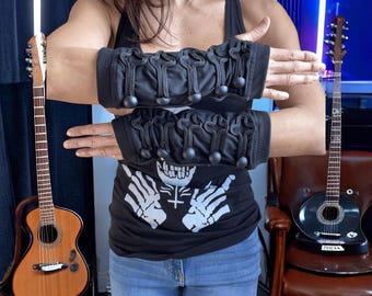 May include: Black arm warmers with decorative black cord and ball accents. The warmers are displayed on a person's arms. A black tank top with a white skeleton graphic is visible. Two guitars are in the background.