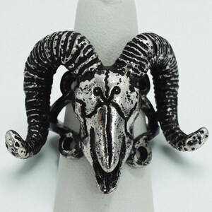 May include: A silver-toned ring featuring a ram skull design. The ring has a detailed, textured finish with prominent horns and a skull-shaped centerpiece. The ring is designed to be worn on a finger, showcasing a gothic or alternative aesthetic.