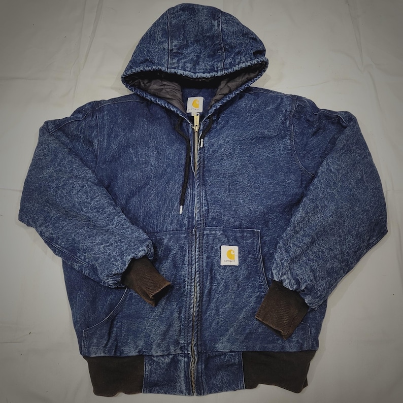 May include: A blue denim hooded jacket with a full zip. The jacket has a quilted lining, ribbed cuffs, and a ribbed hem. The Carhartt logo is on the chest pocket. This workwear-style jacket is designed for warmth and durability, suitable for colder weather.